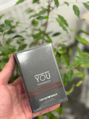 Emporio Armani Stronger With You parfum - Emporio Armani Stronger With You Powerfully är en parfym i en elegant, mörkgrå rektangulär kartong med stilrena bruna linjer och vit text. Förpackningen rymmer 100 ml och utstrålar en modern och exklusiv känsla. Perfekt för dig som gillar lyxiga dofter.