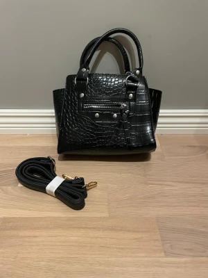 Svart croco handväska med axelrem - Snygg svart handväska i croco-mönstrat konstläder med silverdetaljer och dragkedja framtill. Väskan har både handtag och avtagbar axelrem, samt en rymlig insida med innerfack. Perfekt för dig som vill ha en trendig och stilren accessoar.