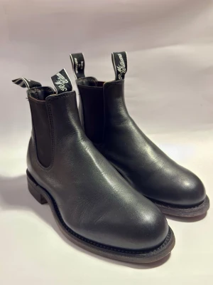 Svarta Chelsea boots från R.M. Williams EU37 - Stilrena svarta Chelsea boots från R.M. Williams i slätt läder. Klassisk rund tå, elastiska sidopaneler och dragflikar med logga upptill. Perfekta för dig som vill ha en tidlös och snygg look med premiumkänsla.