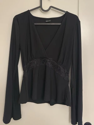 Svart topp från Gina Tricot - Snygg svart omlottopp från Gina Tricot med v-ringning och spetsdetalj under bysten. Långärmad modell i mjukt, stretchigt material som sitter bekvämt och har en feminin siluett. Perfekt till jeans eller kjol för en trendig look.
