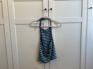 Randig blå linnetopp med knyt - Säljer en snygg randig linnetopp i marinblått, vitt och grönt. Toppen har tunna axelband och knyt i nacken för justerbar passform. Perfekt för varma dagar och enkel att styla med jeans eller kjol.