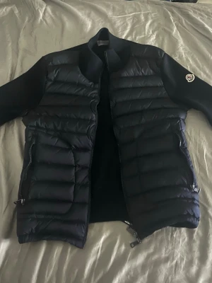 Svart cardigan från Moncler - Snygg svart cardiga nfrån Moncler med quiltad front och släta ärmar. Jackan har dragkedja framtill, två sidofickor med dragkedja och Moncler-logga på ena ärmen. Perfekt för dig som vill ha en stilren och trendig jacka.