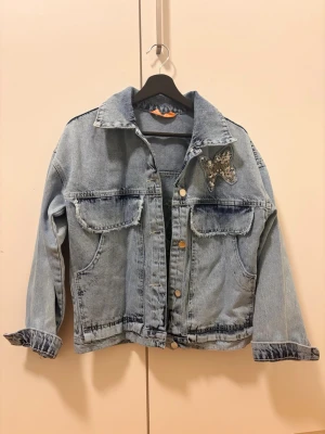 Ljusblå jeansjacka med fjärilsdetalj - Trendig ljusblå jeansjacka med stora fickor framtill och coola knappar. På bröstet och ryggen finns snygga fjärilsapplikationer med paljetter och texten 'Free Flight'. Perfekt för dig som vill sticka ut med en unik denimlook.