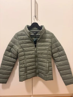 Grön pufferjacka med dragkedja - Snygg grön pufferjacka med hög krage och horisontella sömmar. Jackan har dragkedja framtill och två sidofickor. Perfekt för kalla dagar och ger en stilren look. Materialet är lätt och quiltat för extra värme.