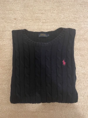 Svart kabelstickad tröja Polo Ralph Lauren - Kabelstickad svart tröja från Polo Ralph Lauren i 100% bomull. Klassisk rund halsringning och rosa broderad logga på bröstet. Perfekt för dig som gillar stilrena och tidlösa plagg med en sportig touch.