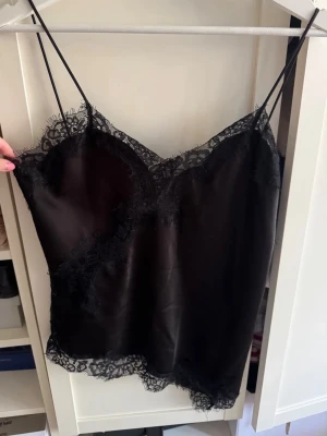 Svart spetstopp  - Snygg svart spetstopp i satinliknande material med tunna axelband och dekorativa spetsdetaljer längs urringning och nederkant. Endast använd vid få tillfällen