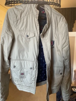 Beige vindjacka från Morris - Stilren beige vindjacka från Morris med ståkrage, tryckknappar och dragkedja. Jackan har två stora fickor framtill, en bröstficka och snygga detaljer som patch med brittisk flagga och Morris-logga på bröstet. Insidan är fodrad med mörkblått mönster.