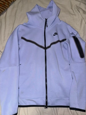 Ljuslila Nike tech - Nike hoodie i ljuslila med hel dragkedja framtill och svart detalj längs bröstet. Har huva, ficka med dragkedja på ärmen och svarta kanter. Tillverkad i mjukt bomullsmaterial, perfekt för en sportig och avslappnad stil.