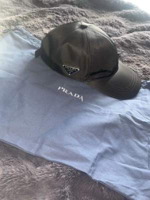 Prada nylon keps - Snygg svart keps från Prada i glansigt nylonmaterial med klassisk böjd skärm och justerbar rem bak. På sidan sitter Pradas ikoniska metalltriangel med logga. Perfekt accessoar för en trendig och sportig look.