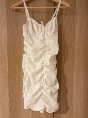 Vit omlott kortklänning med spets - Söt vit kortklänning med smala axelband och omlottdetalj framtill. Klänningen har broderade spetsdetaljer och ett rynkat, figurnära fall. Perfekt för sommarens alla tillfällen och ger en romantisk vibe.