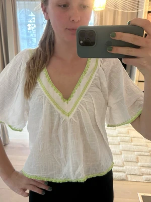 Vit blus med gröna detaljer - Luftig vit blus med v-ringning och ljusgröna broderade kanter. Blusen har korta, vida ärmar och är gjord i ett tunt, krinklat tyg som ger en avslappnad känsla. Perfekt för sommardagar och enkel att styla med jeans eller kjol.