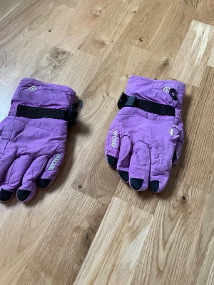 Lila vinterhandskar från Woxx - Säljer ett par lila vinterhandskar från Woxx med Thinsulate-isolering. Handskarna har svarta detaljer på fingertopparna och justerbart band runt handleden. Perfekta för kalla dagar och håller händerna varma.