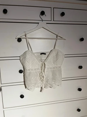 Vit spetslinnetopp med snörning - Supersöt vit linnetopp med tunna axelband, spetsdetaljer och snörning framtill. Toppen har broderade blommor och en v-ringad form. Perfekt för sommarens alla tillfällen och ger en romantisk vibe.