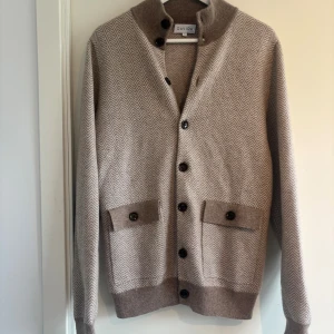 Beige kofta med fiskbensmönster Davida - Snygg beige kofta från Davida med fiskbensmönster och bruna knappar. Koftan har hög krage med extra knäppning, två fickor framtill och ribbade muddar. Perfekt för dig som gillar stilrena och klassiska plagg i mjukt material. Nypris 3499kr, som ny.