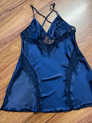 Mörkblått satin nattlinne med spets - Snyggt mörkblått nattlinne i satin med tunna axelband som korsas i ryggen. Dekorativa spetsdetaljer vid bysten och sidorna ger en elegant look. Materialet är glansigt och mjukt, perfekt för en lyxig känsla på kvällen.