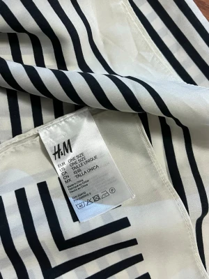 Svartvit randig accessoar från H&M - Snygg sjal från H&M i svart och vitt med grafiskt randigt mönster. Tillverkad i mjuk polyester som känns lätt och följsam. Perfekt accessoar för att lyfta din outfit med en modern touch. One size.