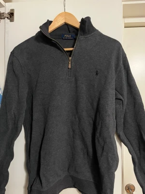 Ralph/Pike - Säljer två snygga halvzip-tröjor från Polo Ralph Lauren. En är mörkgrå och den andra beige, båda med klassisk broderad logga på bröstet. Tröjorna har hög krage, långa ärmar och ribbade muddar. Perfekta för lager-på-lager och stilren vardagslook.