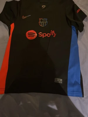 FC Barcelona matchtröja Nike svart - Snygg svart FC Barcelona matchtröja från Nike med Spotify-logga på bröstet och klubbmärke. Tröjan har röda och blå sidopaneler, korta ärmar och trycket 'Lamine Yamal 19' på ryggen. Perfekt för dig som älskar fotboll och vill stötta Barca.
