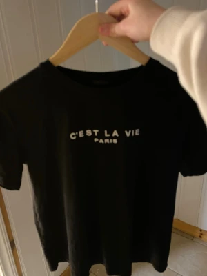 Svart t-shirt med text C'EST LA VIE - Snygg svart t-shirt med vit text 'C'EST LA VIE PARIS' på bröstet. Klassisk rund halsringning och korta ärmar. Perfekt till jeans eller kjol för en chill och trendig look.