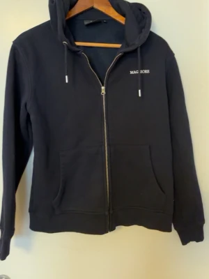 zip hoodie från Maggiore - Mörkblå hoodie med dragkedja från Maggiore. Klassisk modell med huva och dragsnören, samt fickor framtill. Diskret broderad logga på bröstet. Tillverkad i mjukt material som känns skönt mot huden. Perfekt för en avslappnad och stilren look.