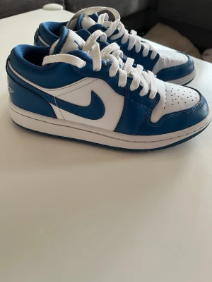 Nike Air Jordan 1 Low blå/vit - Säljer ett par Nike Air Jordan 1 Low sneakers i blått och vitt läder. Klassisk siluett med perforerad tåbox, vita skosnören och ikoniska swoosh-loggan på sidan. Perfekta för dig som gillar streetwear och vill ha en tidlös look.