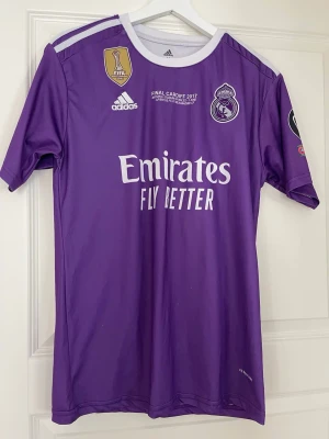 Lila Real Madrid matchtröja Adidas - Säljer en lila Real Madrid matchtröja från Adidas med tryck 'Emirates Fly Better' och Champions League-finalen 2017 på bröstet. Tröjan har vita detaljer, klubbmärke, FIFA-märke och nummer 7 med 'Ronaldo' på ryggen. Perfekt för fotbollsfans! 