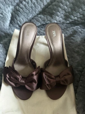 Bruna klackar med rosett från Zara - Snygga bruna klackar från Zara med öppen tå och stor rosett framtill. Skorna har en elegant siluett och är tillverkade i ett glansigt satintyg som ger en lyxig känsla. Perfekta för dig som vill sticka ut med en feminin och trendig detalj. HELT slutsåld på hemsidan