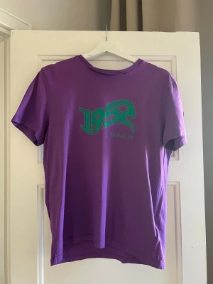 Moncler 1952 t-shirt lila – size M  - Moncler 1952 t-shirt i lila med stor graphic print framtill.  Storlek: M (Moncler size 2) Skick: Bra skick, inga tydliga defekter  Sticker ut med färg och design, perfekt om man vill ha något lite mer unikt. Nypris ca 3000+ kr.  Äkta med QR-verifiering i taggen.  Skriv vid frågor eller för fler bilder