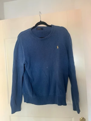 Blå stickad tröja från Polo Ralph Lauren - Klassisk blå stickad tröja från Polo Ralph Lauren med rund halsringning och ribbade muddar. Tröjan har det ikoniska gula broderade logotypen på bröstet och är långärmad. Perfekt för lager på lager och enkel att matcha.