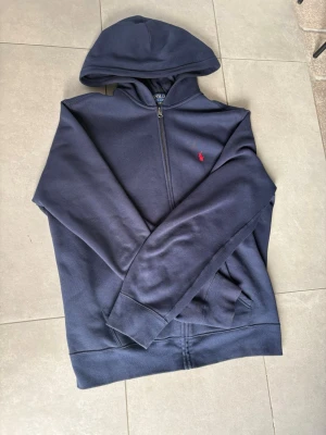 Mörkblå hoodie från Polo Ralph Lauren - Mörkblå hoodie från Polo Ralph Lauren med dragkedja framtill och klassisk röd logga på bröstet. Tillverkad i mjukt bomullsmaterial och har en stor huva samt fickor på sidorna. Perfekt för en avslappnad och stilren look.Är storlek xl men passar som L. Priset kan diskuteras vid snabb äffär!