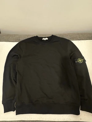 Svart sweatshirt från Stone Island - Svart sweatshirt från Stone Island med klassisk rund hals och ribbade muddar. Ikonisk logotyp-patch på vänster ärm som fästs med knappar. Enkel och stilren design, perfekt för dig som gillar streetwear och vill ha något snyggt och bekvämt.