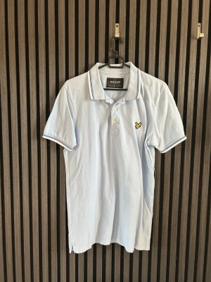Ljusblå pikétröja från Lyle scott - Snygg ljusblå pikétröja från Lyle scott med klassisk krage och knappar framtill. Tröjan har vita och blå detaljer på krage och ärmslut samt en gul broderad logga på bröstet. Perfekt till jeans eller shorts för en avslappnad stil.