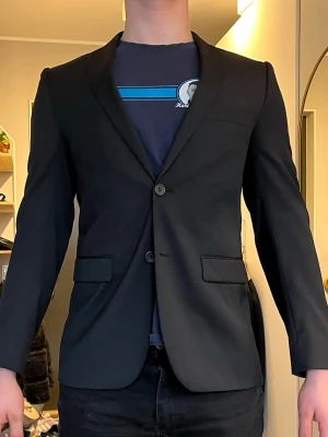 Svart klassisk kavaj herr - Snygg svart kavaj  från dressmann med två knappar framtill och två fickor med lock. Storlek slim fit och regular 46. Figurnära passform och klassisk design som passar till många olika stilar. Hör av dig vid frågor och funderingar!