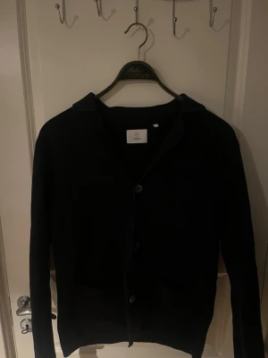 Mörkblå kofta från Bläck med knappar - Stilren mörkblå kofta från Bläck med knäppning framtill och klassisk krage. Perfekt att slänga över en t-shirt eller skjorta för en clean look. Långärmad och mjukt material som känns skönt mot huden.