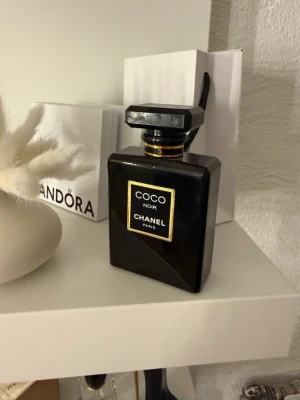 Coco Noir parfym från Chanel - Elegant parfymflaska i svart glas från Chanel, modellen Coco Noir. Flaskan har en fyrkantig form med gulddetaljer och svart etikett. Lock i matchande svart med guldring runt halsen. En lyxig och klassisk doft för dig som gillar exklusiva parfymer.