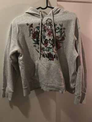 Grå hoodie med blommigt tryck - Säljer en grå hoodie med huva och dragsnören. På bröstet finns ett stort blommigt tryck i vinrött, grönt och lila med texten 'Happiness blooms from within'. Hoodien har en klassisk känguruficka framtill och är gjord i mjukt sweatshirtmaterial. Det står storlek L men den sitter som s och m 