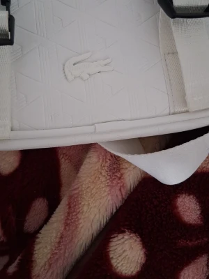 Vit axelväska från Lacoste - Snygg vit axelväska från Lacoste med präglat mönster och den klassiska krokodilloggan på framsidan. Väskan har två svarta spännen, justerbar axelrem och dragkedja upptill. Tillverkad i slitstarkt material med modern, boxig form.