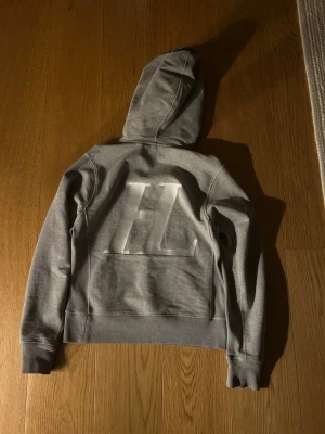 Helmut Lang hoodie - Snygg grå hoodie med stor HL-logga på ryggen och mindre logga på fickan. Klassisk känguruficka framtill och huva med dragsko. Tillverkad i mjukt sweatshirtmaterial, perfekt för chill dagar eller när du vill ha en clean look.