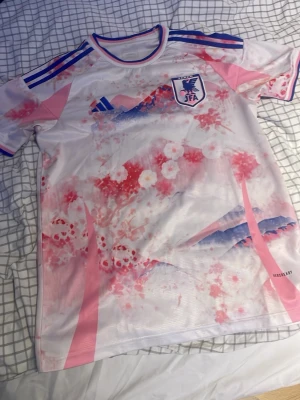 Japan JFA matchtröja rosa/blå Adidas - Säljer en officiell Japan JFA matchtröja från Adidas i vitt med rosa och blå detaljer. Tröjan har ett unikt mönster med körsbärsblommor och berg, samt blå ränder på axlarna. Märkt med JFA-logga på bröstet. Perfekt för fotboll och sport.