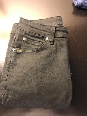 Svarta skinny jeans med stretch - Snygga svarta skinny jeans med klassisk femficksdesign och silverfärgade nitar. Jeansen har en liten gul detalj vid fickan och en svart läderpatch bak i midjan. Materialet är stretchigt för extra komfort och passform. Perfekta till en avslappnad streetstyle-look.