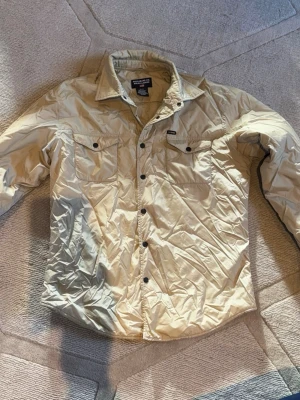 Beige overshirt från Ralph Lauren - Stilren beige overshirt från Ralph Lauren i glansigt material. Två bröstfickor med knappar och klassisk krage. Perfekt att bära öppen eller stängd för en avslappnad look. Svarta tryckknappar framtill och diskret logga ovanför fickan.