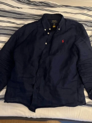 Marinblå skjorta från Polo Ralph Lauren - Klassisk marinblå skjorta från Polo Ralph Lauren med knappkrage och den ikoniska röda logotypen broderad på bröstet. Skjortan har långa ärmar och knappar framtill. Tillverkad i bomull för en skön och stilren känsla.