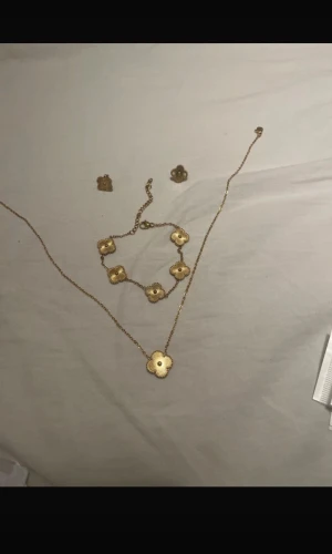 Guldfärgat smyckeset med blommor - Säljer ett smyckeset i guldfärg med matchande halsband, armband och örhängen. Alla delar har en söt blomformad design med detaljerade kanter och en liten kula i mitten. Perfekt för dig som gillar unika accessoarer med blommotiv.