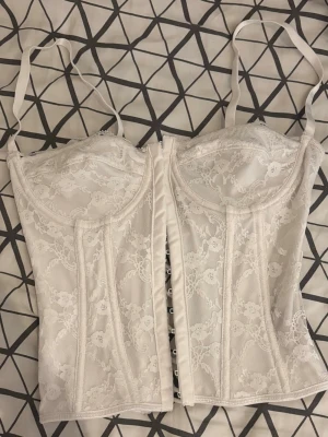 Vit spetskorsett med snörning - Säljer en elegant vit korsett med tunna axelband och vacker spetsdetalj över hela plagget. Perfekt för dig som vill ha en feminin och stilren look.