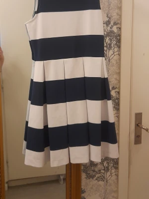 Randig klänning från Polo Ralph Lauren - Snygg ärmlös klänning från Polo Ralph Lauren med breda marinblå och vita ränder. Klänningen har en utställd kjol med veck och rund halsringning. Tillverkad i mjuk bomull som känns skön mot huden. Perfekt för en fräsch och stilren look.