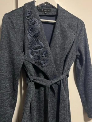Mörkgrå kappa med broderi från Alvina - Stilren mörkgrå kappa från Alvina med bälte i midjan och unika broderade blommor med pärldetaljer på ena slaget. Kappan har lång ärm och mjukt, stickat material som ger en avslappnad men elegant look. Väldigt fin men kommer tyvvär inte till användning , priset går att diskutera  passar M/S /Xs 