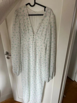 Adoore klänning  - Långklänning i vit med små blå blommor, v-ringad framtill och med långa ärmar. Klänningen har en mjuk, flytande passform och knäppning i ryggen. Perfekt för sommarens alla tillfällen.