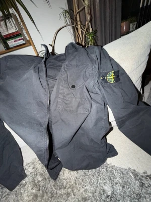 Svart vindjacka från Stone Island - Snygg svart vindjacka från Stone Island med dragkedja framtill och en bröstficka med knapp. Klassisk patch med logga på vänster ärm. Lätt material som passar perfekt till streetwear och vardagsstil. Perfekt skick, XL men sitter som L