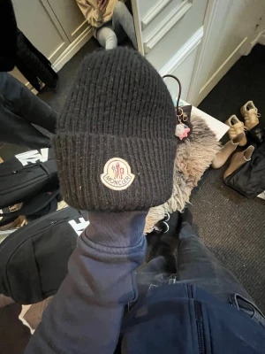 Svart ribbstickad Moncler mössa - Svart ribbstickad mössa från Moncler med uppvikt kant och klassisk Moncler-logga framtill. Mössan är tillverkad i ett mjukt och värmande material, perfekt för kalla dagar. Snygg och enkel design som passar till de flesta outfits.