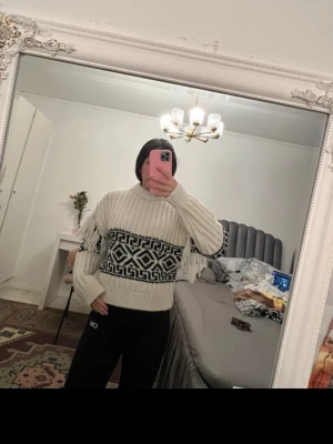 Stickad tröja med mönster och fransar - Säljer en beige stickad tröja med svartvitt grafiskt mönster runt bröstet och ryggen. Tröjan har fransar längs sidorna, ribbstickade muddar och hög hals. Perfekt för dig som gillar unika detaljer och mysig stil.
